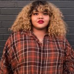 a Black woman with curly blonde hair that's dark at the roots stands facing the camera while wearing an orange and brown plaid button-down