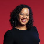 color photograph headshot of a femme person with shoulder-length curly black hair wearing gold hoop earrings and glasses. She smiles at the camera and wears a black blouse with slightly puffed shoulders against a red background